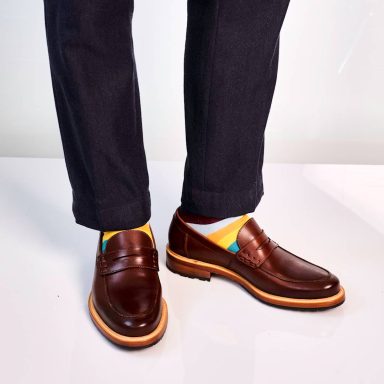 Braune Mokassins mit orangefarbener Sohle und bunten Socken, getragen mit dunkelblauen Hosen.