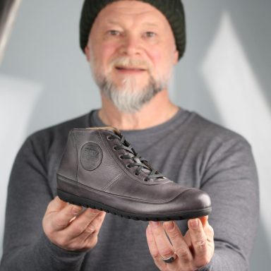 Mann hält einen grauen Stiefel in der Hand, lächelnd, mit Mütze und grauem Shirt.
